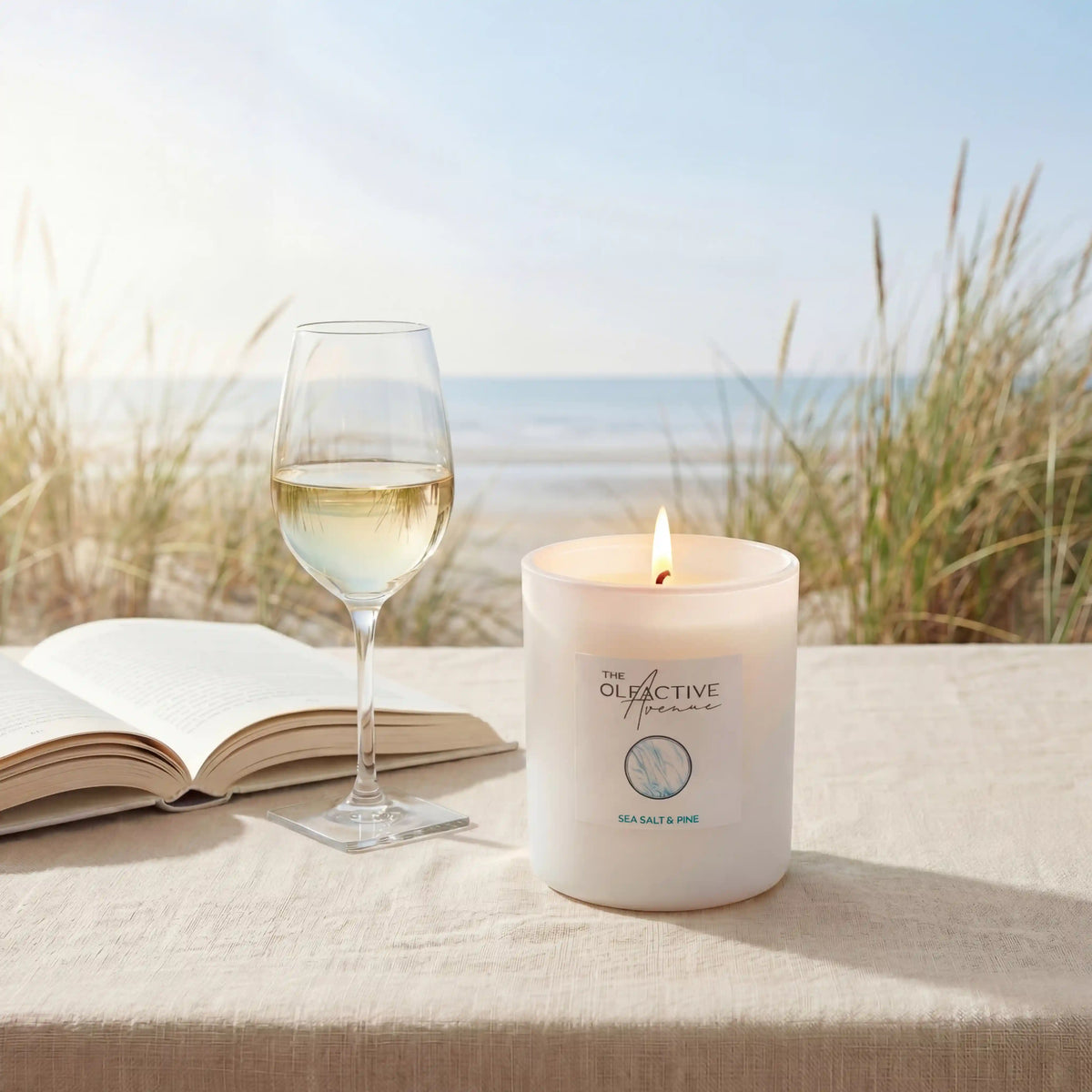 Duftkerze Sea Salt & Pine von The Olfactive Avenue am Strand mit Weinglas und Buch – Atmosphère Frisch & Klar.