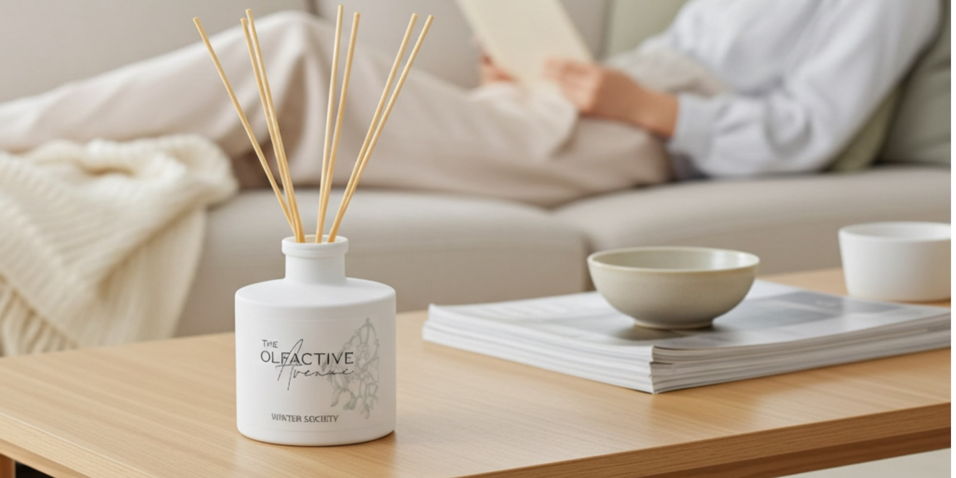 Cozy Wohnzimmer: Stäbchen-Diffusor von The Olfactive Avenue auf Couchtisch, im Hintergrund Person lesend auf dem Sofa
