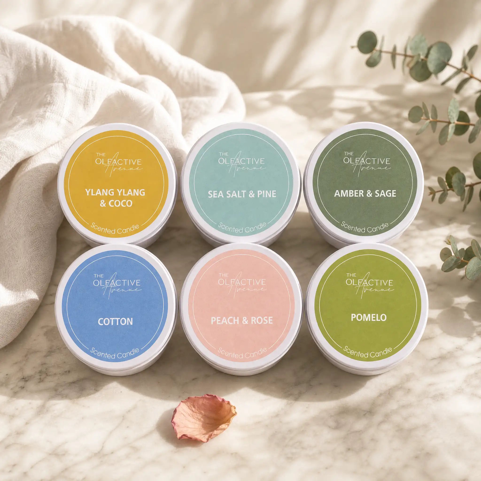 Hochwertiges Travel Candle Bundle von The Olfactive Avenue mit sechs kleinen Duftkerzen auf hellem Marmor, Leinenstoff und Eukalyptus. Das Bundle kombiniert florale, frische, cremige und aromatische Duftnoten für stilvolle Duftmomente zu Hause oder unterwegs.