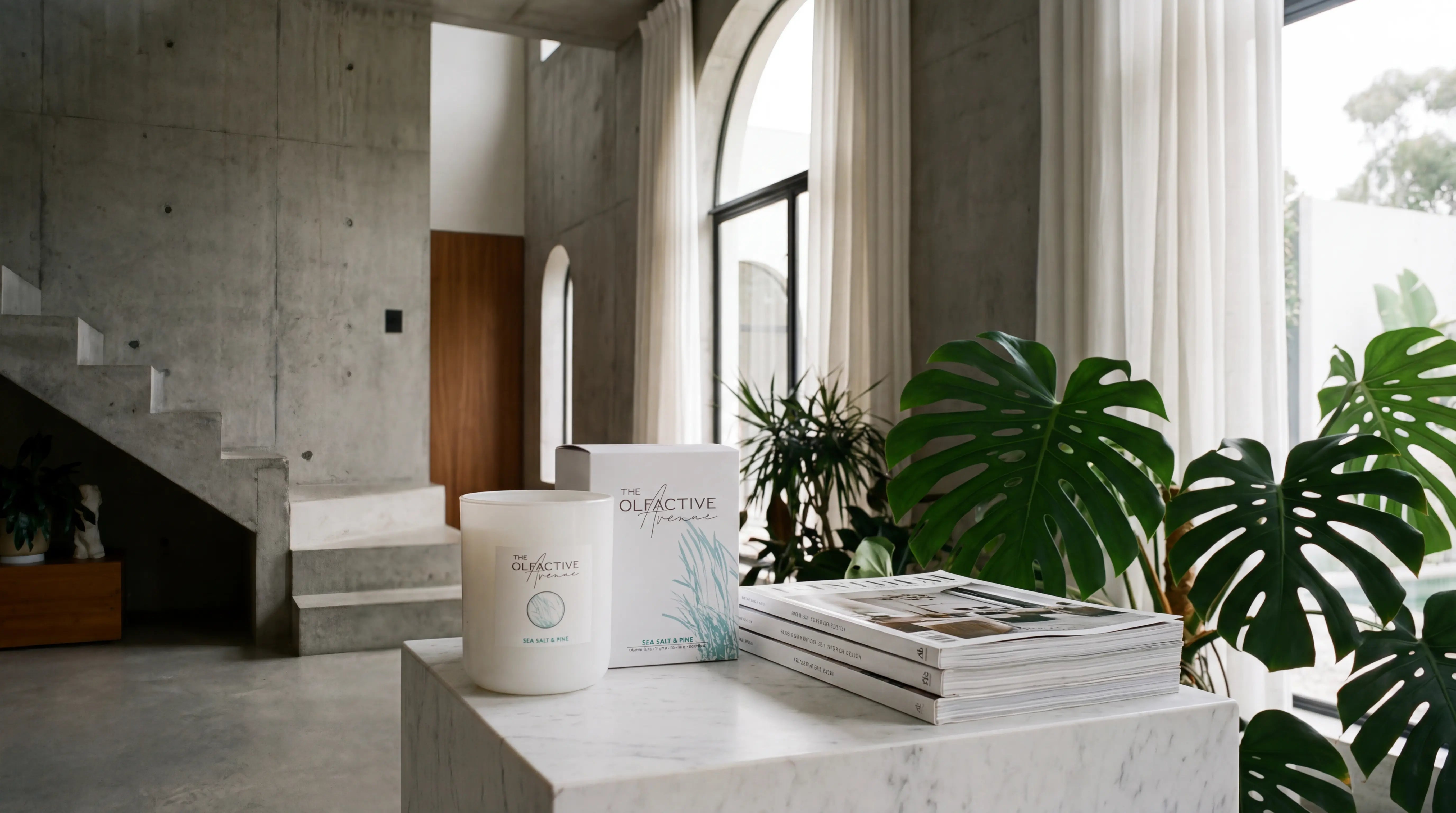Luxuriöse, matte weiße Duftkerze und Verpackung von The Olfactive Avenue aus pflanzlichem Raps- und Kokoswachs, arrangiert auf einem Marmor-Sockel neben einem Stapel Magazine und einer großen Monstera-Pflanze in einem sonnendurchfluteten, minimalistischen Interieur.