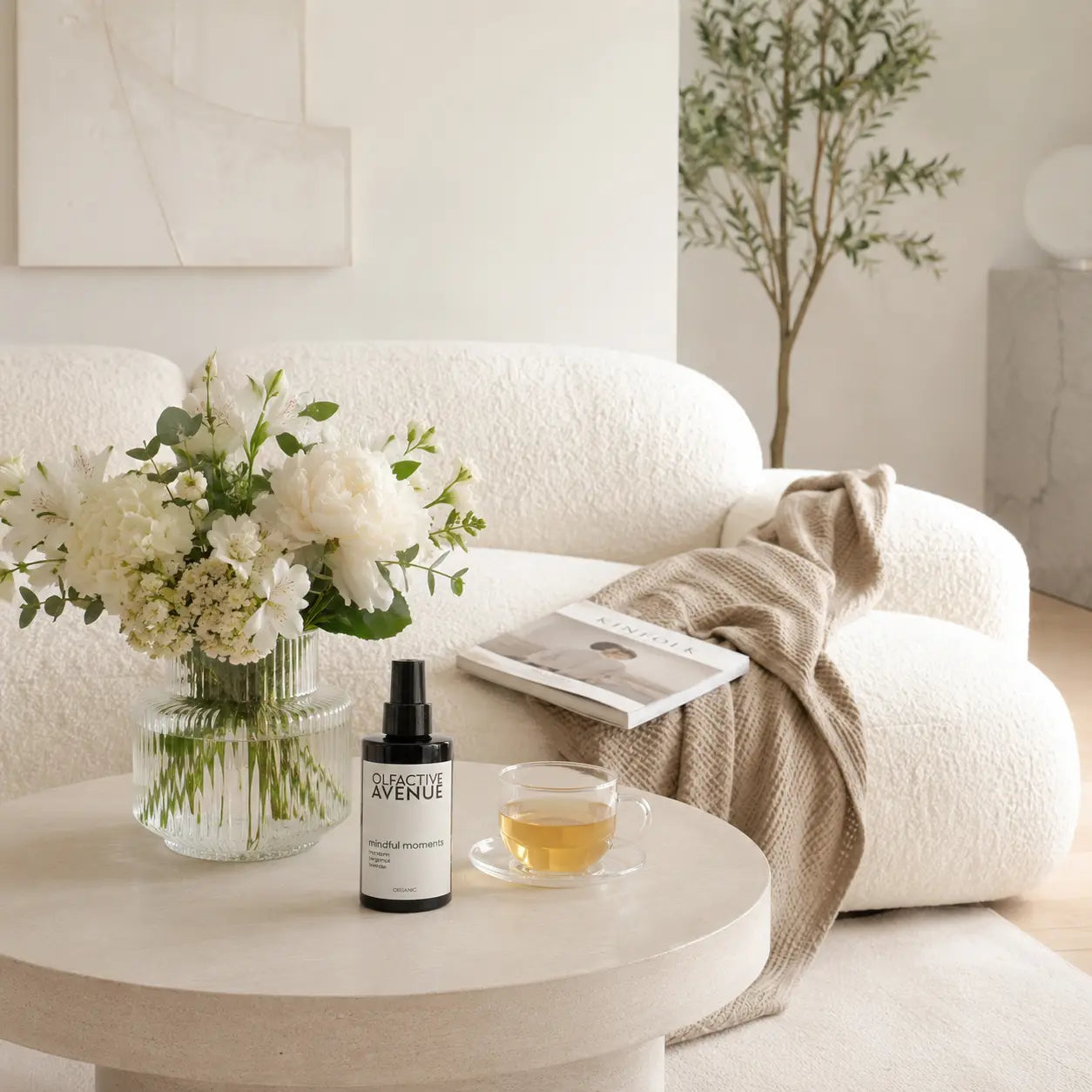 Mindful Moments Bio-Raumspray von The Olfactive Avenue auf einem Couchtisch mit Tee, Blumen und hellem Wohnzimmer-Ambiente.