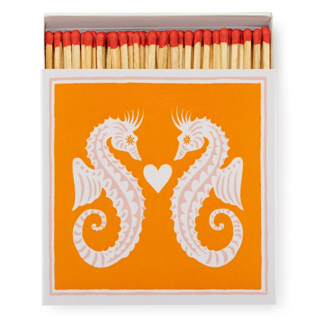 Quadratische Matchbox „Seahorses“: zwei weiße Seepferdchen mit Herz auf Orange; 125 lange Streichhölzer, natur mit roten Köpfen, weißes Tray.