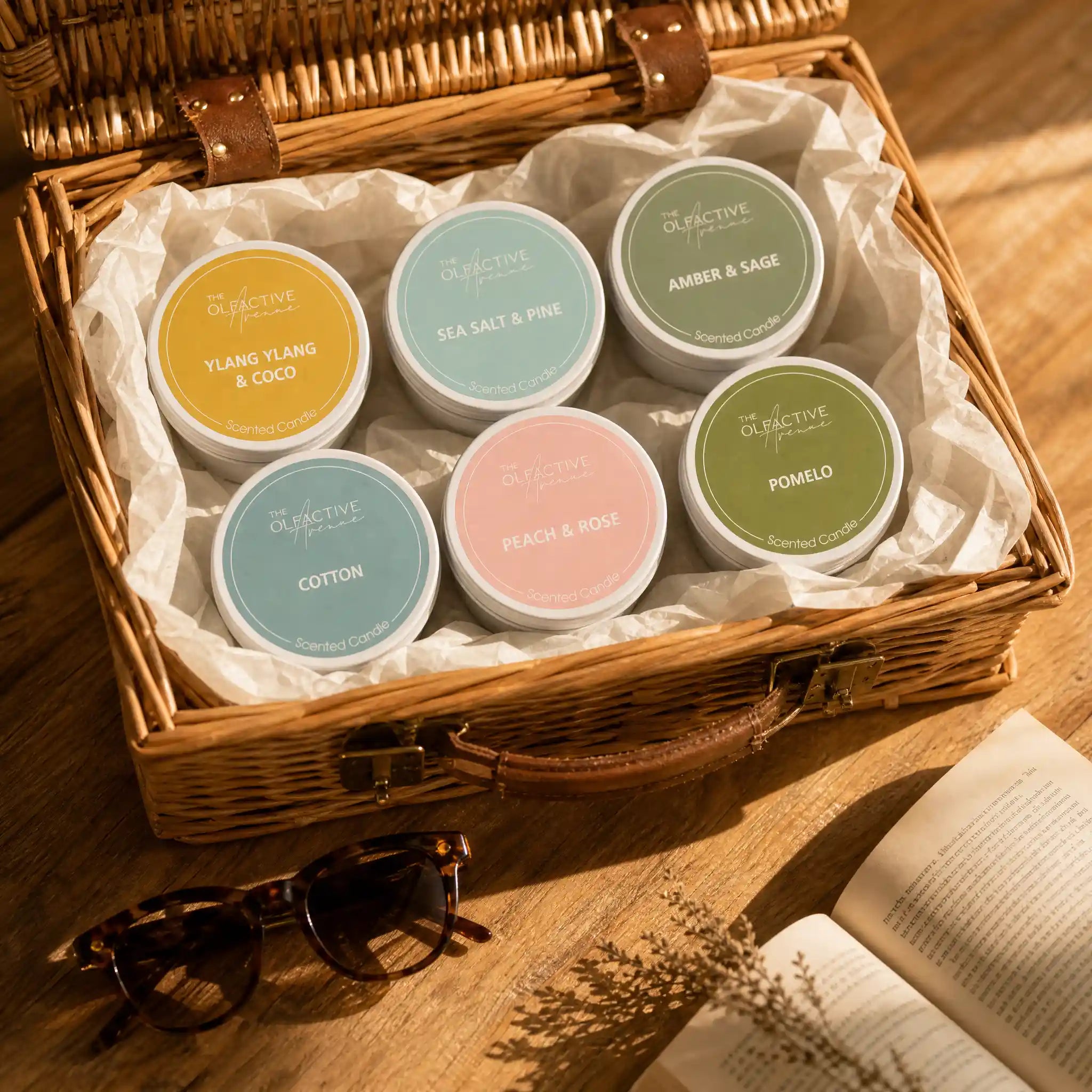 Sechs Travel Candles von The Olfactive Avenue in einem geflochtenen Reisekorb mit Seidenpapier, Sonnenbrille und Buch im warmen Reise-Lifestyle-Setting.