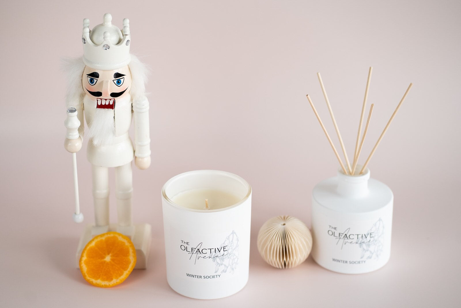 Hero-Bild der Winter Edition: The Olfactive Avenue „Winter Society“ als Duftkerze und Raumduft-Diffuser, puristisches Setting mit Nussknacker und Orangenscheibe.