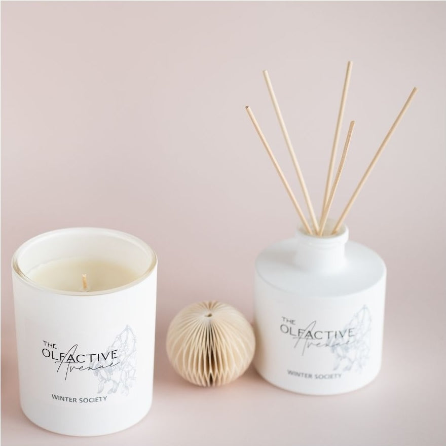 Quadratisches Produktbild: The Olfactive Avenue „Winter Society“ als Duftkerze und Raumduft-Diffuser in puristischem Setting auf warmem Rosé-Hintergrund.