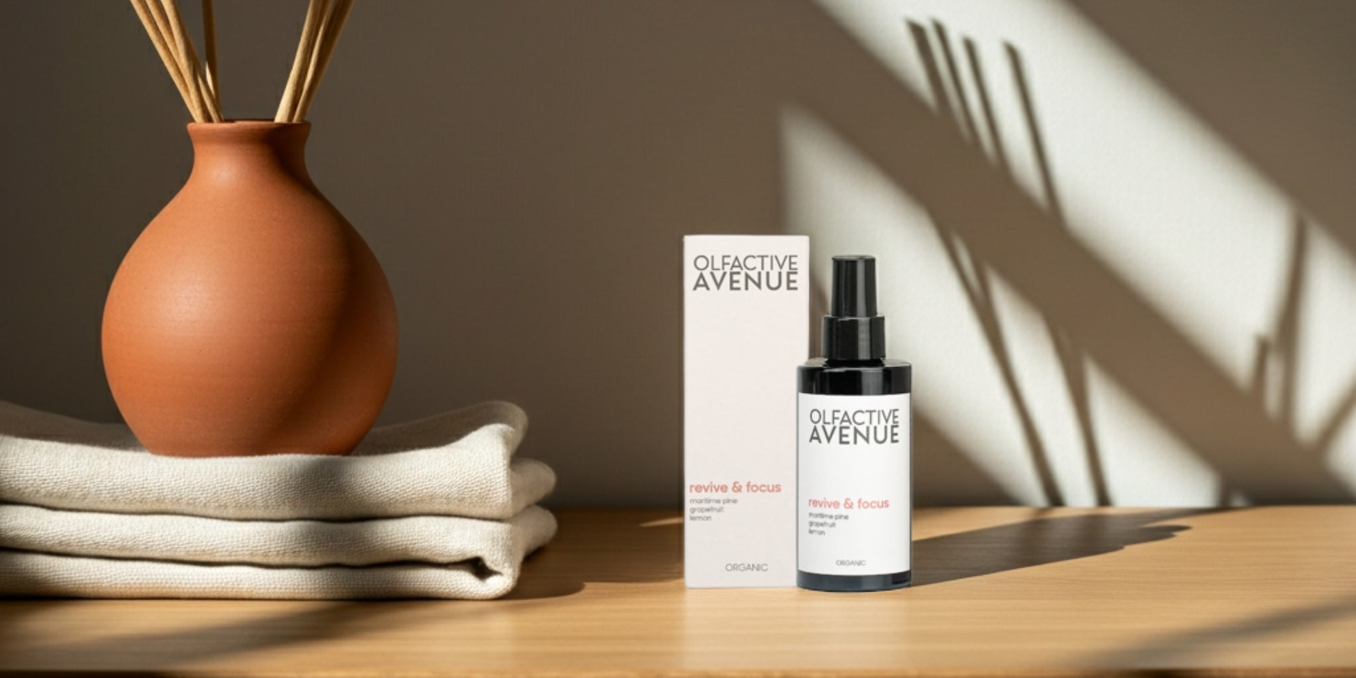 Bio-Raumspray ‘revive & focus’ von The Olfactive Avenue mit Verpackung, Tonvase und Leinen auf Holz, weiches Abendlicht.